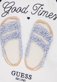 Gráfico de dos sandalias con correas texturizadas de color azul claro y suelas beige. Se incluyen los textos "Buenos Momentos" y "GUESS EST. 1981".