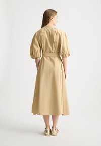 Beiges Midikleid mit Puffärmeln, taillierter Taille und fließendem Rock. Aus leichter Stoffqualität. Die Rückansicht zeigt einen Gürtel und Falten.