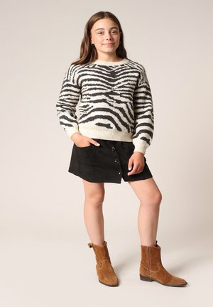 Pull à motifs zèbre en crème et noir, associé à une jupe ensuède noire avec des boutons. Des bottines en suède marron complètent la tenue.
