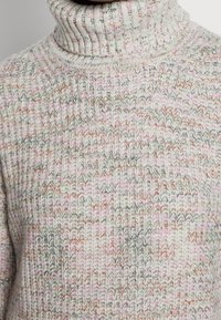 Maglione a collo alto lavorato a maglia in grigio chiaro con accenti rosa, arancione e verde. Tessuto texturizzato con schemi di cucitura visibili.