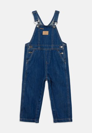 Salopette en denim bleu foncé avec bretelles ajustables, boutons en métal sur les côtés, poches avant et petite pièce carrée en cuir sur la poitrine.