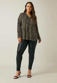 Schwarze Bluse mit langen Ärmeln und einem strukturierten Muster aus goldenen Sprenkeln, kombiniert mit glänzenden schwarzen Leggings und schwarzen Riemchen-Heels.