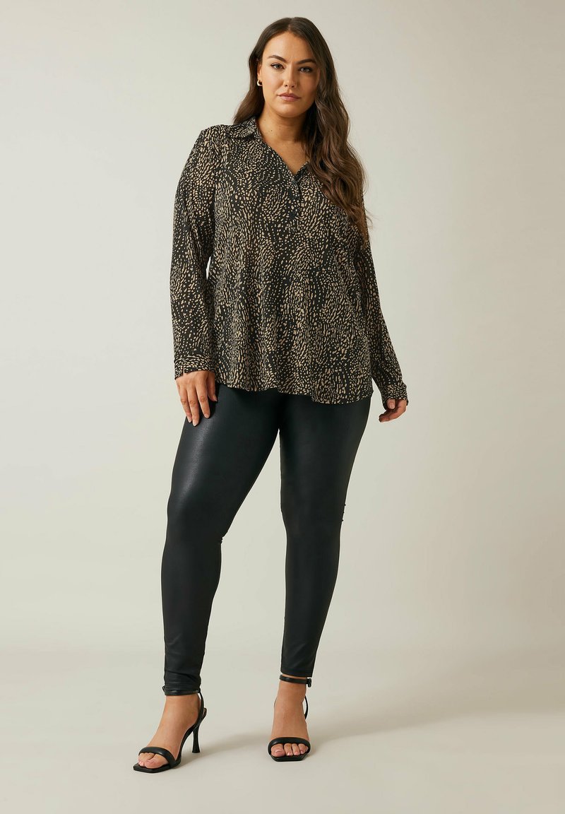 Schwarze Bluse mit langen Ärmeln und einem strukturierten Muster aus goldenen Sprenkeln, kombiniert mit glänzenden schwarzen Leggings und schwarzen Riemchen-Heels.