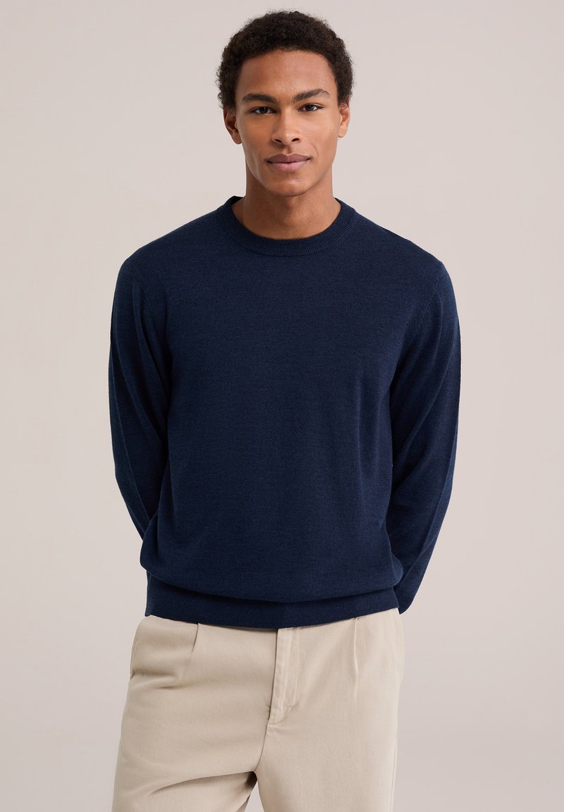 Jeune homme avec des cheveux courts bouclés portant un pull à manches longues bleu marine et un pantalon beige, debout devant un fond uni.