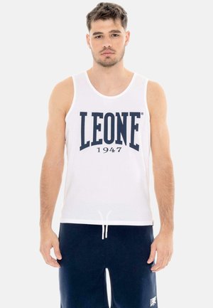 Homme portant un haut blanc sans manches avec le texte « LEONE 1947 » et un pantalon bleu marine, debout devant un fond clair uni.
