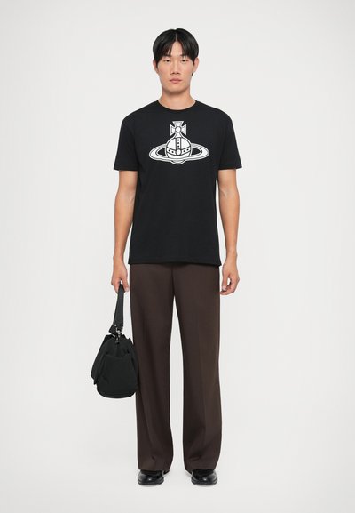 T-shirt en coton noir avec un design graphique blanc, associé à un pantalon large marron et un sac en tissu noir.