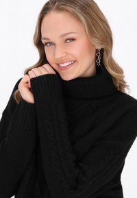 Schwarzer, gestrickter Rollkragenpullover mit strukturiertem Muster. Verfügt über gerippte Bündchen und eine lockere Passform. Ein Paar goldene Kettenohrringe sichtbar.