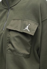 Veste vert olive avec un col zippé et une poche poitrine arborant un logo blanc de basketball Jumpman sur le rabat.