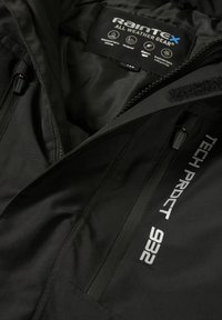 Veste noire imperméable avec poche zippée étiquetée "TECH PRDCT 928" et une étiquette intérieure montrant les caractéristiques de l'équipement toutes saisons RainTex.