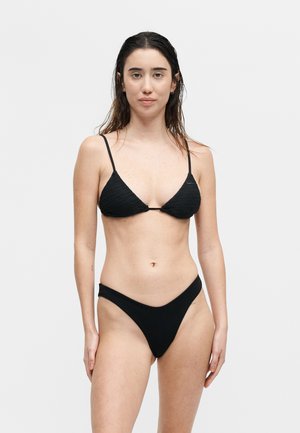 Femme aux cheveux mouillés portant un bikini texturé noir avec un haut triangle et un bas taille haute, debout devant un fond clair uni.