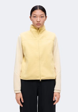 Jeune femme aux cheveux foncés porte un gilet polaire jaune pâle par-dessus une chemise crème à manches longues et un pantalon noir, debout devant un fond uni.