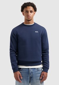 Jongeman met krullend haar draagt een navyblauwe sweatshirt met het logo "Ballin" en lichtblauwe jeans, staande tegen een effen lichtgrijze achtergrond.