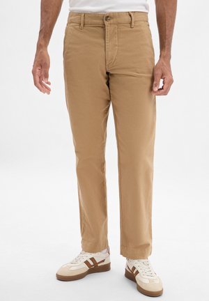 Beige Baumwollhosen mit geradem Schnitt, ausgestattet mit einem Frontknopfverschluss und seitlichen Taschen, kombiniert mit weißen und braunen Sneakers.
