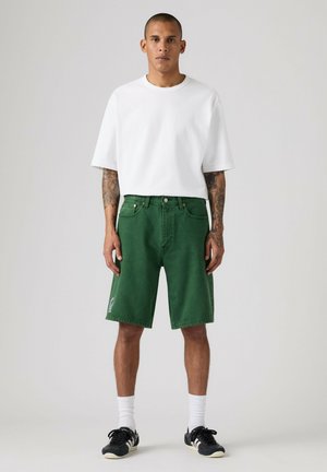 Man met tatoeages die een effen wit T-shirt, groene knielange korte broek, witte sokken en zwarte sneakers draagt, staand tegen een effen achtergrond.