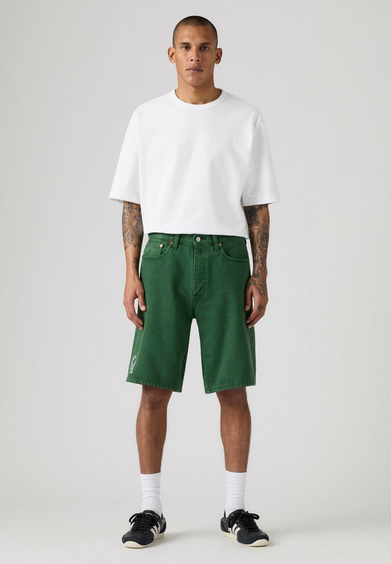 Mann mit Tätowierungen, der ein schlichtes weißes T-Shirt, grüne kniehohe Shorts, weiße Socken und schwarze Turnschuhe trägt, steht vor einem einfarbigen Hintergrund.