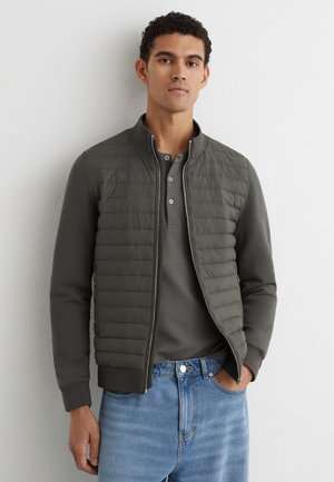 Jonge man draagt een donkergrijze doorgestikte jas met ritssluiting over een grijze henley-shirt en lichtblauwe spijkerbroek, staand tegen een witte muur.