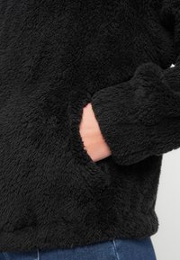 Μαύρο μπουφάν από φλίς με υφή, που διαθέτει πλαϊνή τσέπη και απαλό, βελούδινο άγγιγμα. Τα μανίκια είναι ελαφρώς γυρισμένα στις μανσέτες.