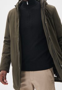 Man i en olivgrön jacka över en svart kabelstickad tröja med dragkedja och beige byxor, stående med armarna något uppsträckta.