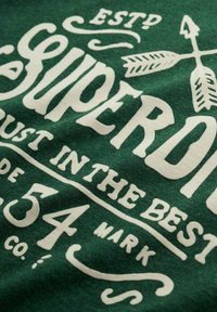 Grøn stof med hvid tekst i vintage-stil, der fremhæver pile og dekorative elementer, med fokus på brandnavnet og forskellige sætninger.