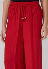 Pantalons rouges, légers, amples avec une taille élastique et un cordon de serrage, featuring des accents en bois et des poches latérales.