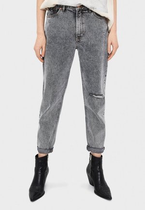 Jean en denim gris à jambes droites, avec des revers roulés et un détail usé au genou gauche. Associé à des bottines noires.