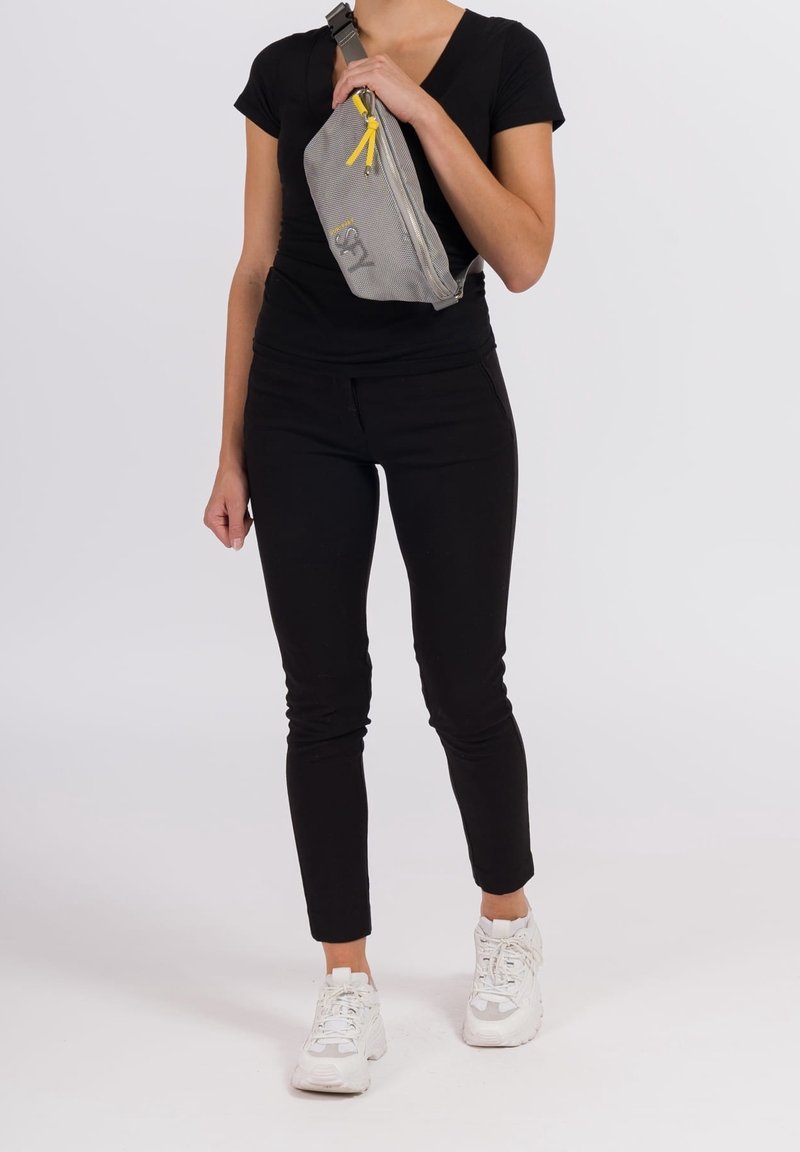 Personne portant un t-shirt noir à manches courtes, un pantalon noir, des baskets blanches, et tenant un sac bandoulière gris avec des fermetures éclair jaunes.
