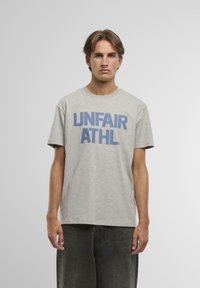 Šedé bavlněné tričko s krátkým rukávem, na přední straně se středovým umístěním se nachází výrazný modrý text "UNFAIR ATHL". Normální střih, kulatý výstřih.