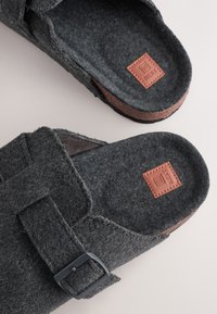 Chaussures grises en feutre à enfiler avec une boucle noire, une semelle intérieure en liège marron contrastante et une étiquette logo en cuir embossé sur la semelle intérieure.