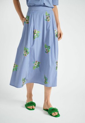 Midi rok met blauwe en witte strepen en geborduurde kleurrijke bloemmotieven, gedragen met groene, textuurige slip-on sandalen.