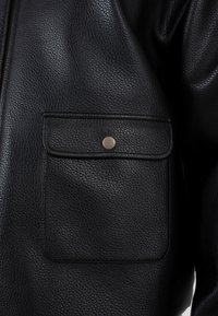 Detailní záběr kapsy na černé kožené bundě s texturou, která má klopu zajištěnou jediným kovovým knoflíkem.