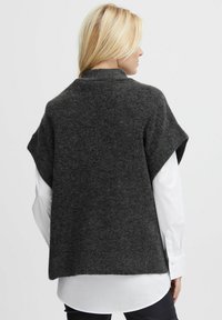 Grau gestrickter Poncho mit kurzen Ärmeln, weicher Textur, gerundetem Halsausschnitt und lockerer Passform, über einem weißen Hemd getragen.