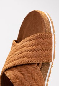 Sandali marroni con texture, caratterizzati da un motivo intrecciato sulle cinghie, dotati di un sottopiede in sughero e una suola in gomma bianca.