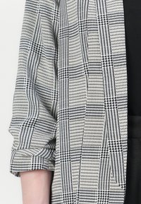 Černobílý kostkovaný blazer s texturovanou vazbou, s ohrnutými rukávy a štíhlým střihem. Černá látka pod ním.