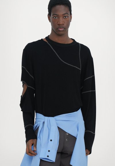 Schwarzes Langarmshirt mit grauen Nähdetails und Cut-out-Schultern, kombiniert mit einem hellblauen, um die Taille gebundenen Shirt.