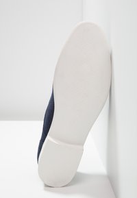 Semelle d'une chaussure bleu marine inclinée contre un angle blanc, montrant une semelle extérieure en caoutchouc blanc lisse avec des détails de grip légèrement texturés.