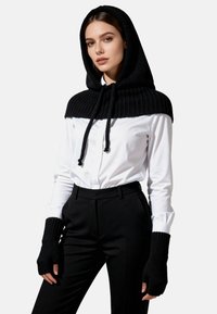 Cappa con cappuccio lavorata a maglia nera con texture a coste, dotata di coulisse regolabile; abbinata a una camicia bianca con bottoni e pantaloni neri su misura.