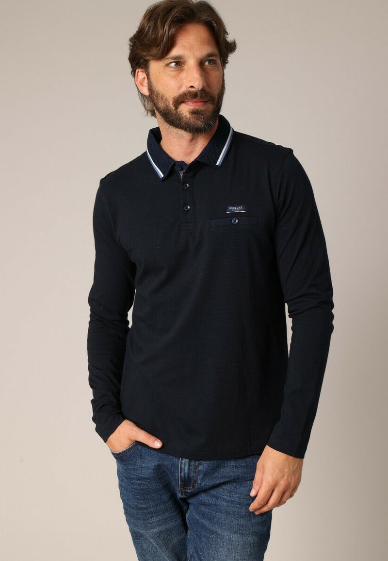 Polo à manches longues en bleu marine avec un col blanc contrastant, une patte de boutonnage à deux boutons et une poche sur la poitrine. Le tissu semble lisse et texturé.