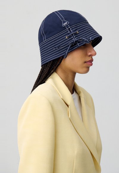 Cappello a secchiello blu navy con cuciture bianche, dotato di lacci laterali e un'etichetta con il marchio. Indossato con un blazer giallo chiaro.