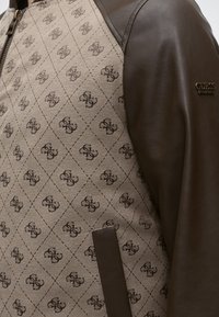 Tissu beige à motif avec impression répétée de logo marron, associé à une manche en cuir marron foncé et une étiquette de marque en métal Guess sur le haut du bras.
