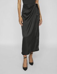 Gonna avvolgente nera in satin con dettaglio annodato sul lato, lunghezza fino al polpaccio, texture liscia, abbinata a tacchi a punta neri.