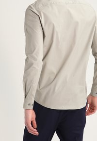 Man med en ljusbeige långärmad knappad skjorta och mörkblå byxor, sedd bakifrån med avslappnade armar vid sidorna.