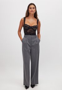 Body in pizzo nero con scollatura a cuore, abbinato a pantaloni grigi a vita alta e plissettati. Vestibilità sartoriale e texture liscia.