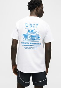 Άνδρας που φοράει άσπρο T-shirt με μπλε γραφικό ενός κτιρίου και κείμενο που γράφει «OBEY House of Subversion» και ονόματα πόλεων στην πλάτη.