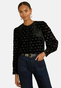 Melna blūze ar sirds rakstu, garām caurspīdīgām piedurknēm un apaļu kakla izgriezumu. Apvienota ar tumši zilu džinsu biksēm un melnu jostu ar zelta detaļām.