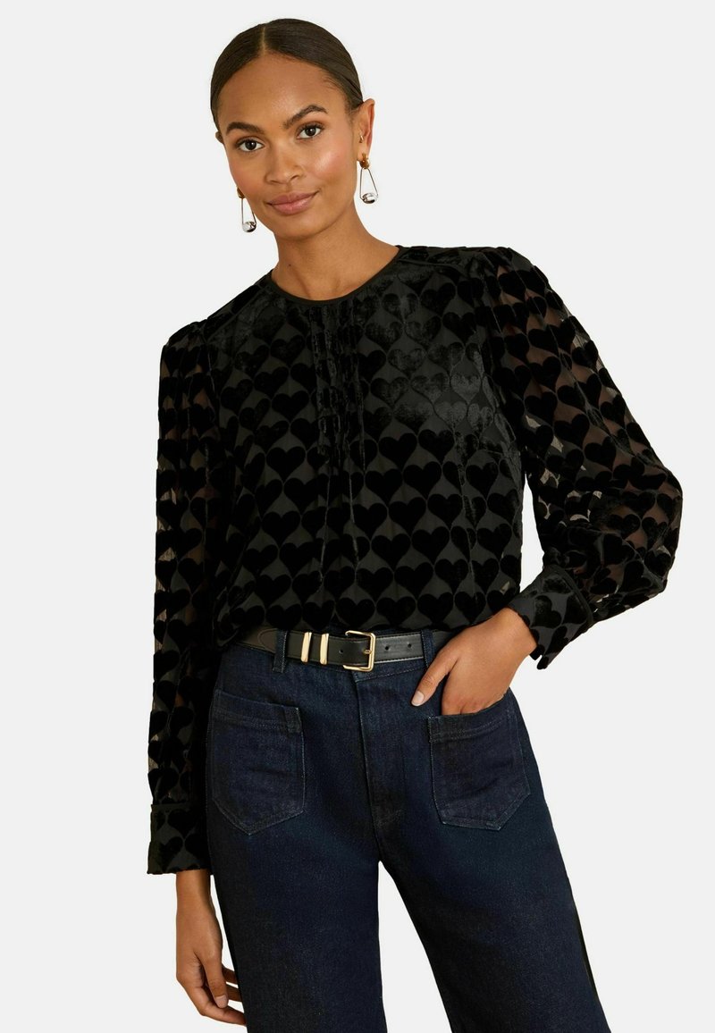 Melna blūze ar sirds rakstu, garām caurspīdīgām piedurknēm un apaļu kakla izgriezumu. Apvienota ar tumši zilu džinsu biksēm un melnu jostu ar zelta detaļām.