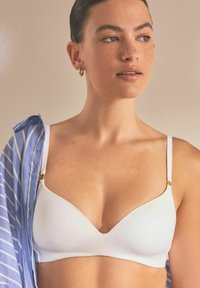 Witte, gevormde bralette met smalle bandjes en gouden hardwareaccenten, gecombineerd met een blauw gestreept shirt. Soepele stof en een V-vormige halslijn.