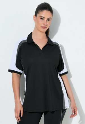 Mujer con cabello oscuro recogido en una cola de caballo, vistiendo una camiseta deportiva de manga corta en blanco y negro y leggings negros sobre un fondo blanco.