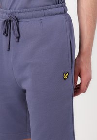 Violetinių medvilnės mišinio šortų su elastingu juosmuo, raišteliu, šoninėmis kišenėmis ir geltonu išsiuvinėtu logotipu kairėje kojoje.
