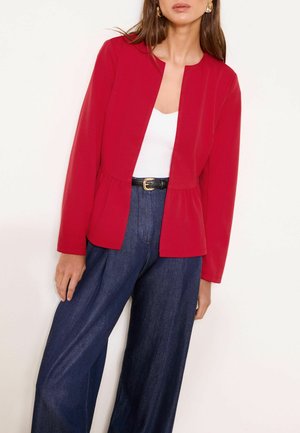 Frau trägt eine rote Jacke mit offenem Vorderteil, weißes Oberteil, hoch taillierte dunkelblaue weite Hosen und einen schwarzen Gürtel mit goldener Schnalle.