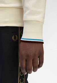 Felpa crema con polsini a coste caratterizzati da righe blu e bordeaux, abbinata a pantaloni neri decorati con ricami dorati a alloro.